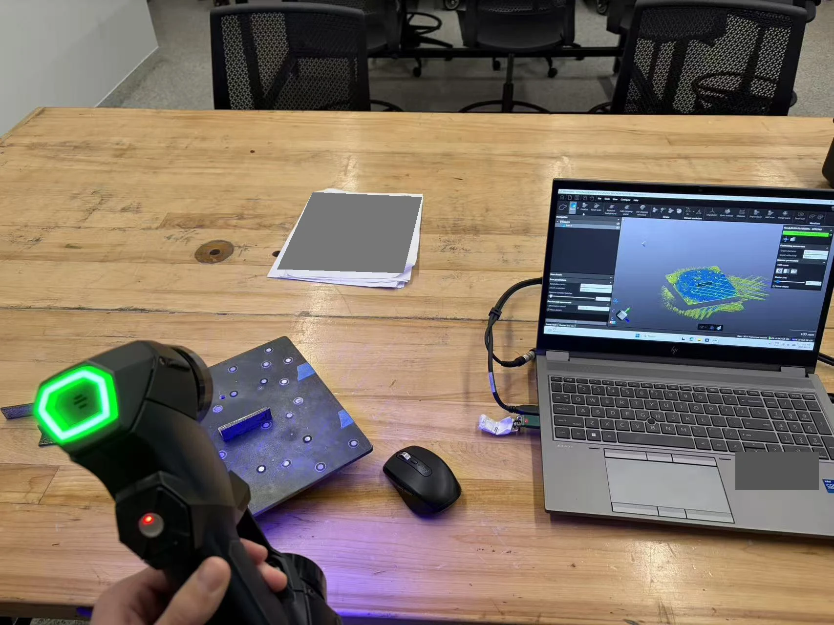 Handheld 3D scanner capturing a metal specimen beside a laptop displaying scanned geometry